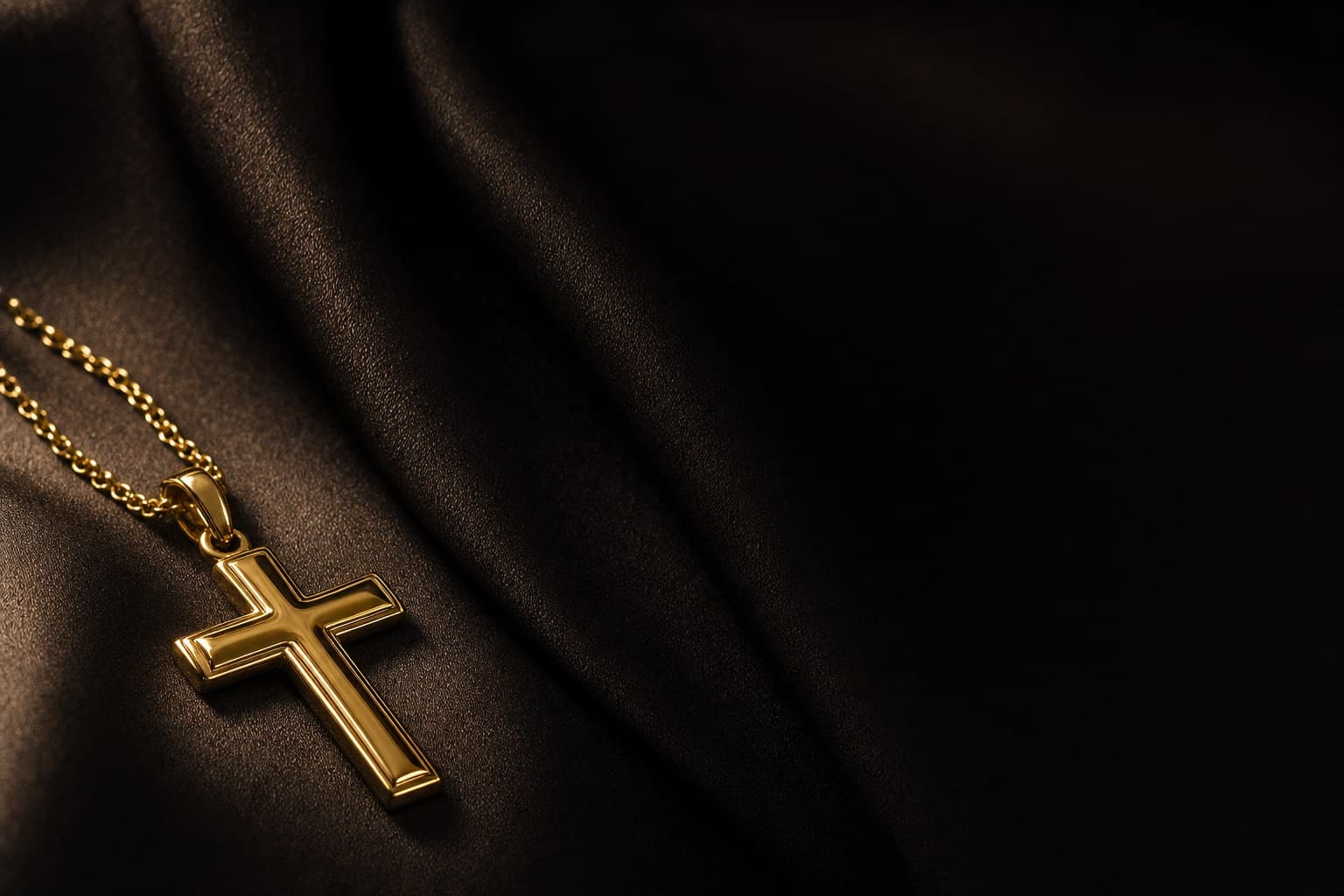 A cross in 18K yellow gold on a fine chain, resting on deep charcoal silk in raking light