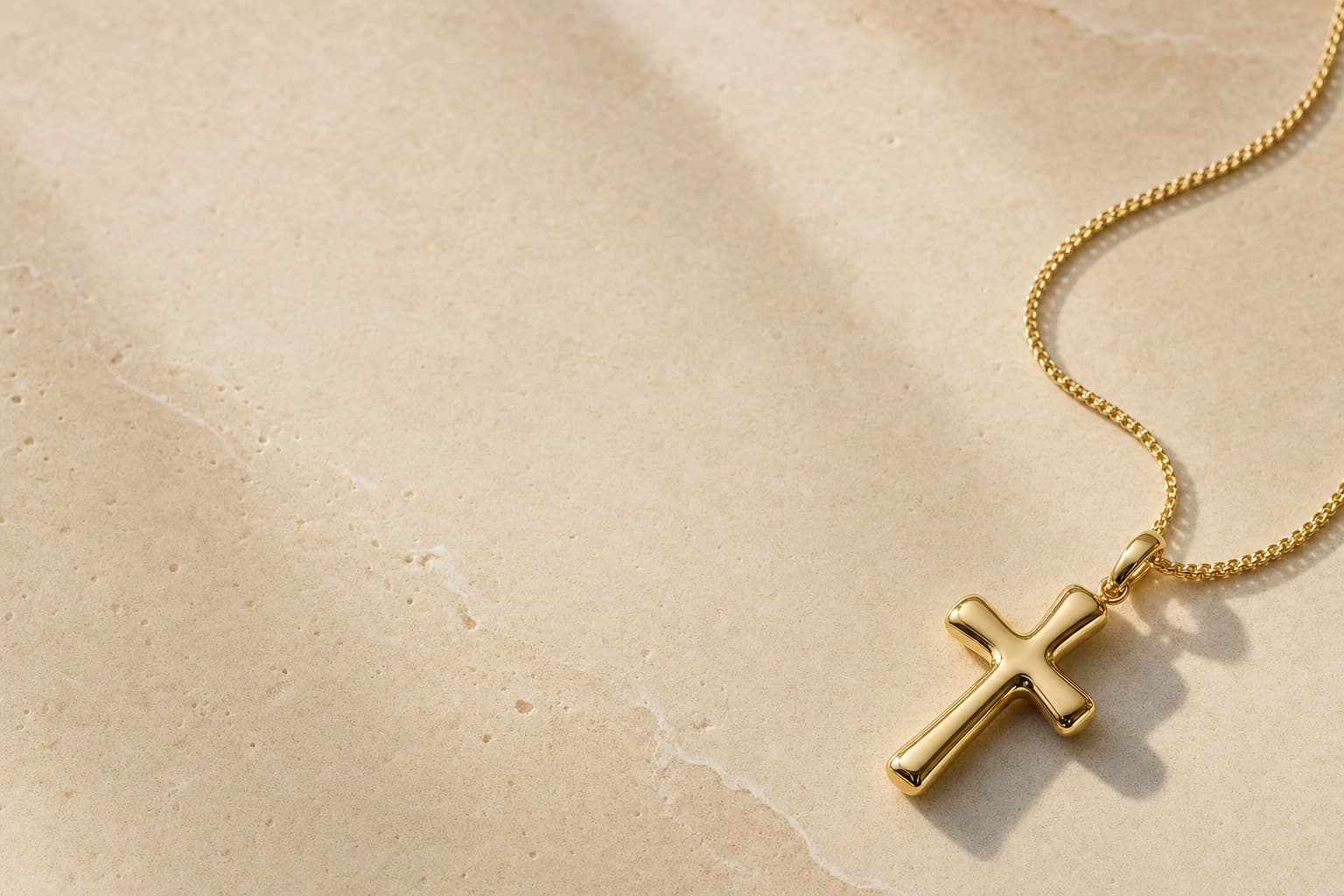 A cross in 18K yellow gold on a fine chain, resting on warm ivory travertine in soft daylight